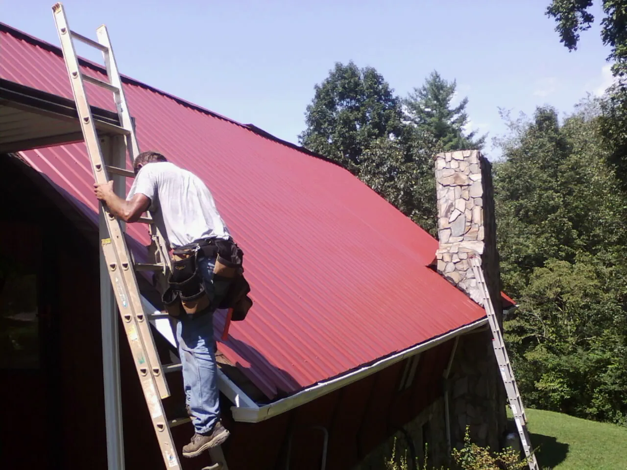Roofer on a ladder beside a red metal roof and stone chimney — Soffit & Fascia Repair by Roofing Brothers in Kaw City